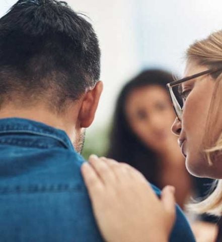 woman comforting a man wearing a blue denim shirt by placing a hand on his shoulder